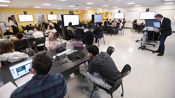 A professor prepares his lesson at the podium while students log into their laptops